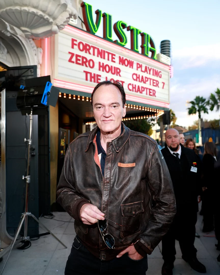 Ünlü yönetmen Quentin Tarantino, üzerinde A. Tarantino yazan kahverengi yıpranmış deri ceketiyle, sağ elinde güneş gözlüğünü tutarken, arkasında neon ışıklı VISTA Sineması tabelasında Fortnite Now Playing Zero Hour: The Lost Chapter yazısının görüldüğü bir etkinliğin girişinde kameraya bakıyor.