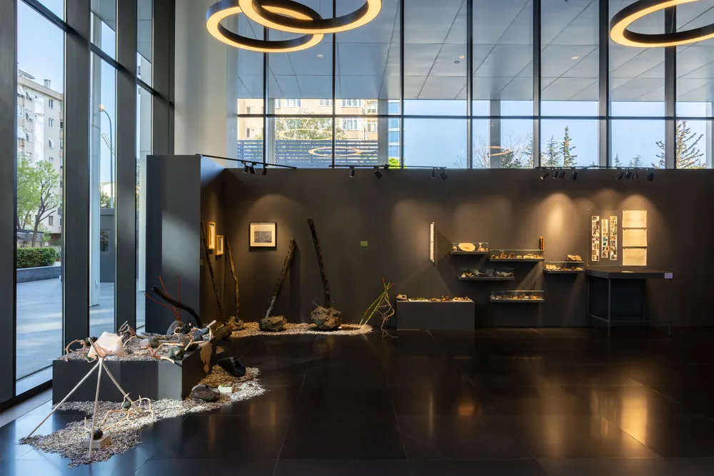 Quick Art Space sergisinden geniş bir iç mekan, karanlık gri duvarlar boyunca spot ışıklarıyla aydınlatılmış çeşitli sanat eserleri, vitrinlerdeki küçük objeler ve zeminde doğal malzemelerden oluşan büyük bir enstalasyon sergileniyor. Geniş cam pencerelerden dışarıdaki şehir manzarası ve tavandaki halka şeklindeki aydınlatmalar modern bir atmosfer yaratıyor.