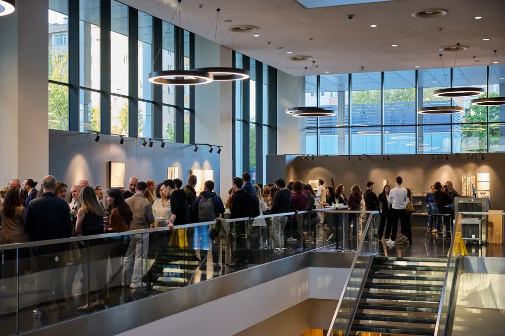 Modern ve geniş bir iç mekanda, duvardaki sanat eserlerinin önünde kalabalık bir grup, Quick Art Space sergi etkinliğinde sohbet edip eserleri incelerken, tavandan sarkan halka şeklindeki şık aydınlatmalar ve büyük cam pencereler mekanı aydınlatıyor.