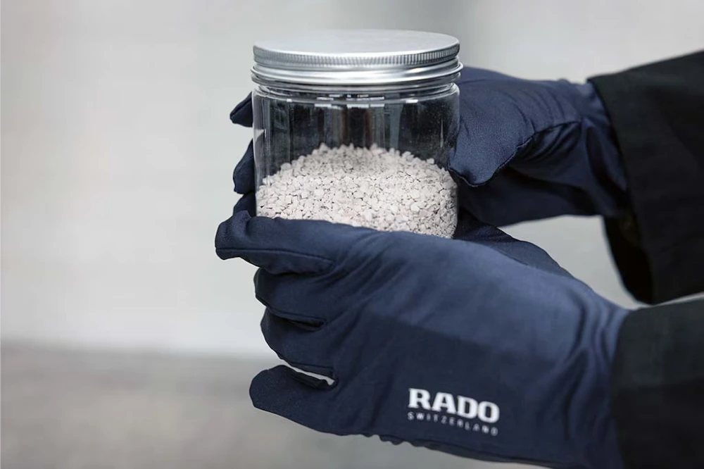 Rado markalı koyu renk eldivenler giyen bir kişinin elleri, markanın ileri teknoloji seramik saatlerinin üretiminde kullanılan, açık renkli ve granül formdaki zirkonyum oksit tozunu barındıran şeffaf bir cam kavanozu özenle tutuyor.