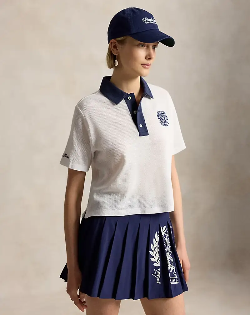 Koyu lacivert Wimbledon şapkası, lacivert yakalı ve patlı, göğsünde işlemeli Wimbledon amblemi olan Ralph Lauren pamuklu polo tişörtü ve beyaz desenli lacivert pilili tenis eteği giyen zarif bir kadın model, hafif sağa dönük poz veriyor.