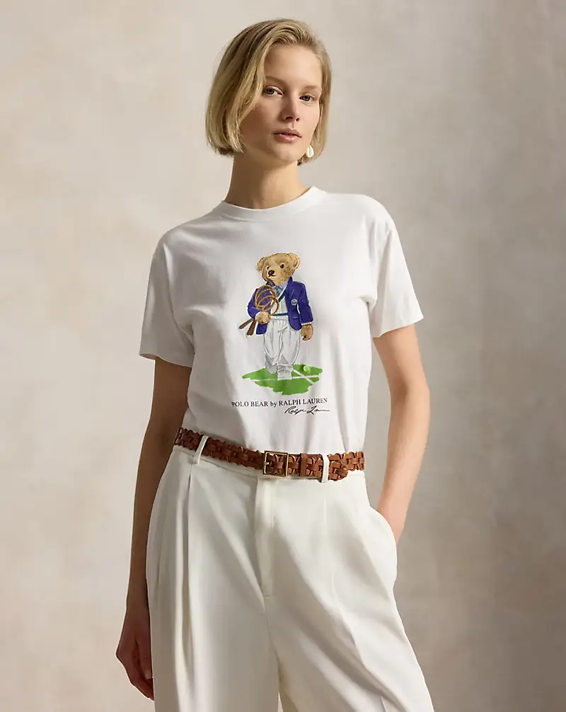 Sarışın, kısa saçlı bir kadın, üzerinde tenis raketi tutan ve blazer ceket giyen bir Polo Bear illüstrasyonunun bulunduğu Ralph Lauren Wimbledon temalı beyaz bir tişörtü, beyaz bol paça pantolon ve kahverengi örgülü bir kemerle kombinleyerek poz veriyor.