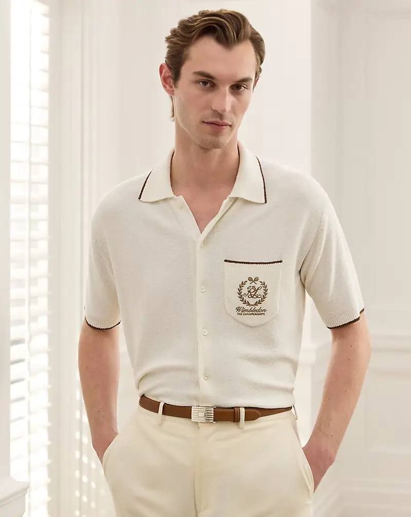 Krem rengi, yakalı ve düğmeli, kısa kollu triko hırka giyen, sol göğüs cebinde işlenmiş Wimbledon logosu ile yaka ve kol ağzında kahverengi biye detayları bulunan şık bir genç adam, aynı tonda pantolon ve kahverengi kemerle kombinini tamamlayarak kameraya bakıyor.