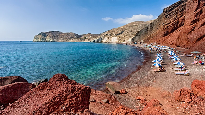 Santorini Kırmızı Plajı'nda, güneşli bir günde, kıyıya dizilmiş çok sayıda mavi-beyaz şemsiyenin altında uzanan tatilcilerin, berrak turkuaz sularla çevrili kızıl kayalıkların ve kumların oluşturduğu eşsiz manzaranın keyfini çıkardığı görülüyor.