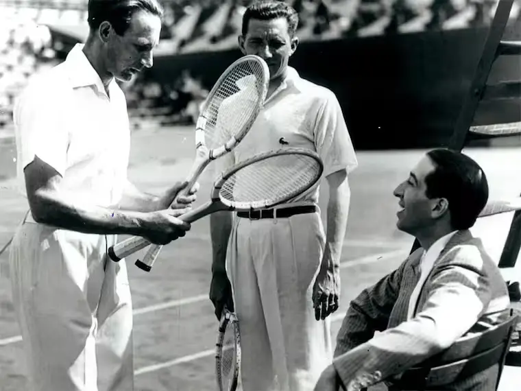 Siyah beyaz bu fotoğrafta, bir tenis kortunda, ortada timsah logolu tişörtüyle tanınan René Lacoste'un ayakta elinde raketle durduğu, solunda başka bir tenisçinin iki raketi dikkatle incelediği ve sağda sandalyede oturan takım elbiseli bir beyefendinin gülümsediği, tarihten bir tenis anını yakalıyor.