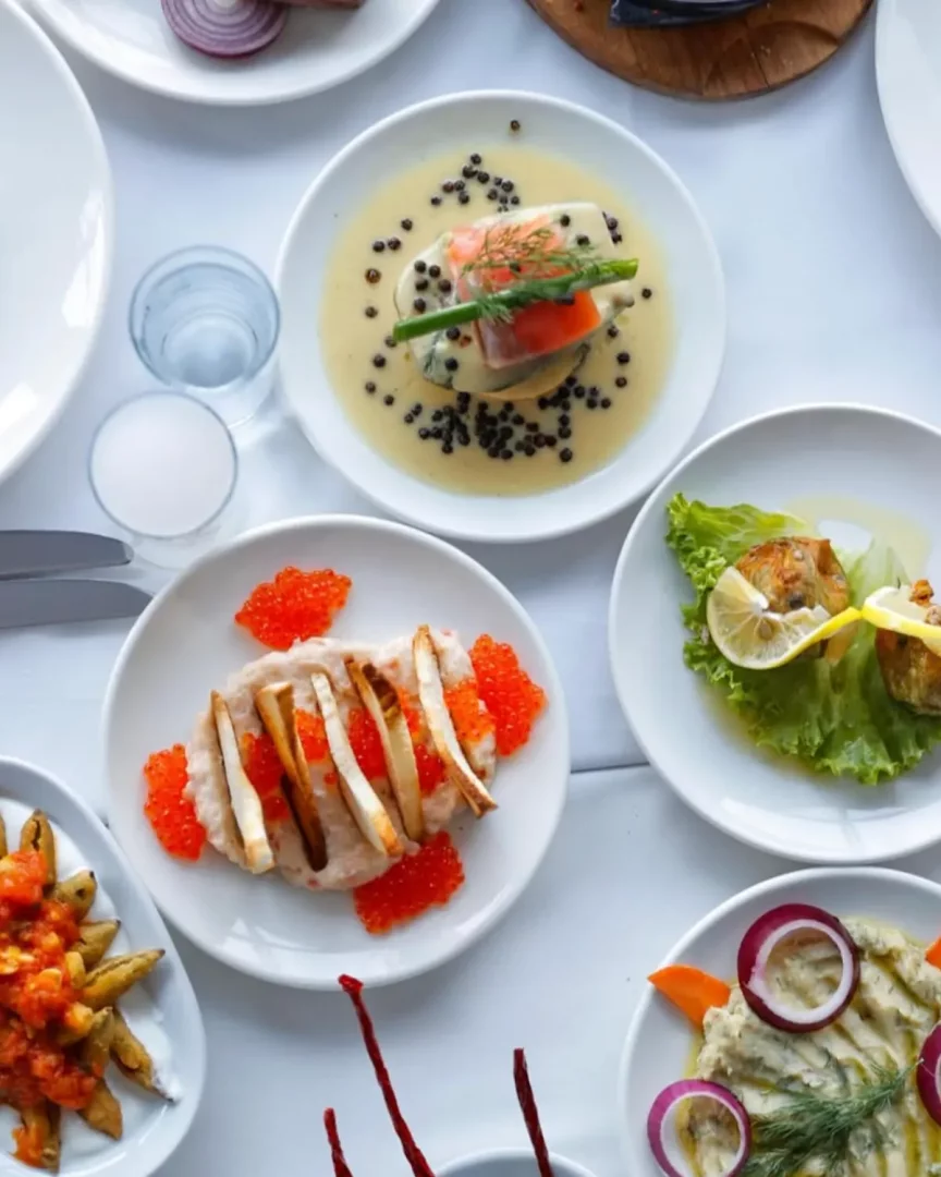 Arnavutköy'deki bir balık restoranında beyaz bir masanın üzerinde sunulan, kremalı somon, parlak kırmızı havyarla süslenmiş fava, limon dilimli kızarmış balık ve mor soğanlı mezeler gibi özenle hazırlanmış çeşitli deniz ürünleri ve meze tabakları.