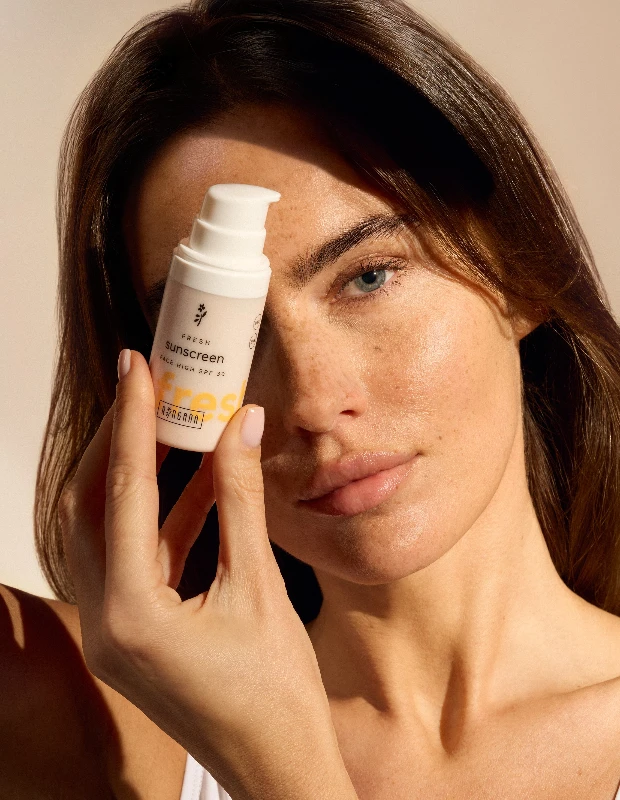 Yüzünü kısmen kapatacak şekilde elinde FRESH sunscreen FACE HIGH SPF 30 yazılı bir güneş kremi şişesi tutan bir kadın, berrak mavi gözleri ve doğal çilleriyle sağlıklı cilt için taze kozmetik ve güneş korumasının önemini vurguluyor.