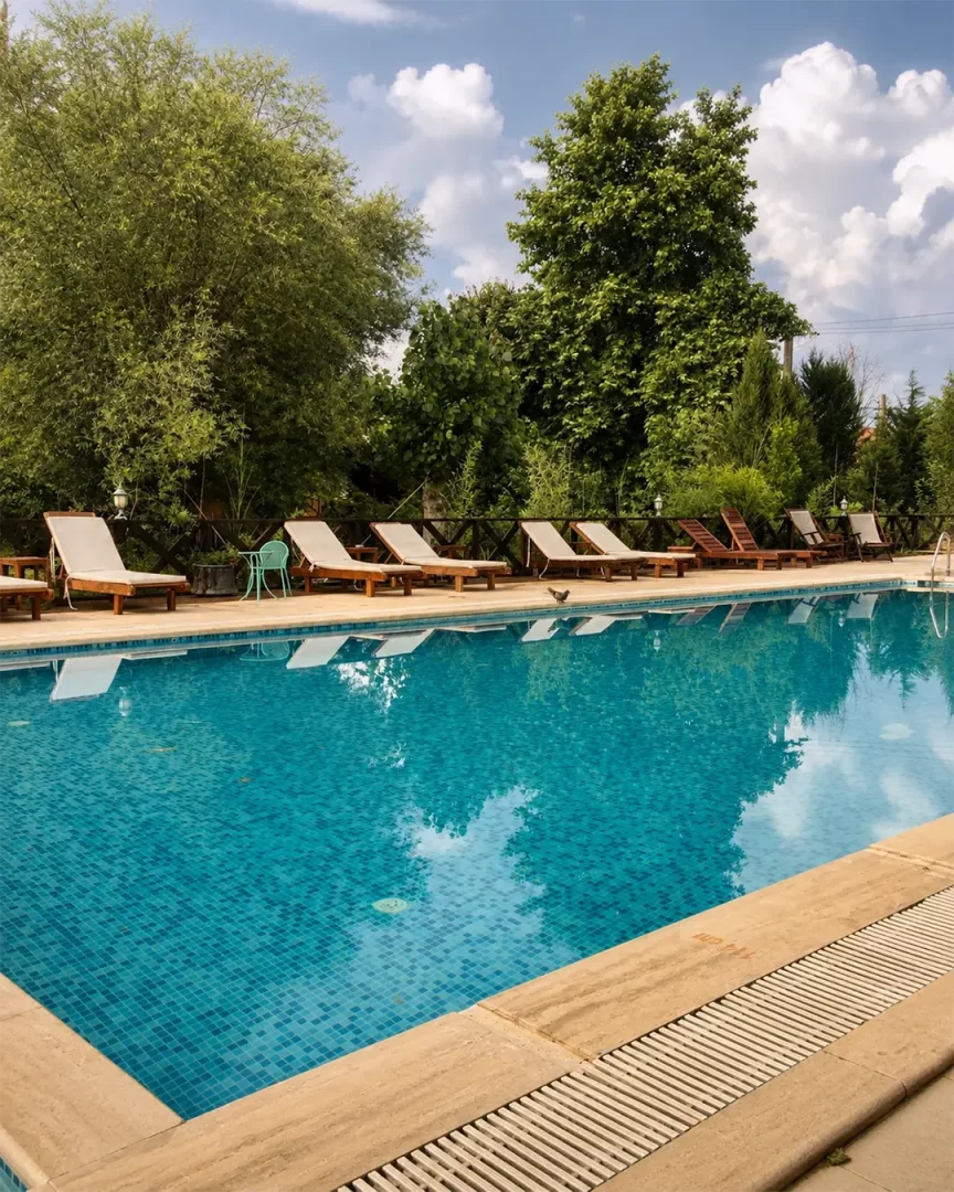 Robins Nest Eco Resort'taki bu canlı fotoğraf, berrak mavi mozaik karolarla kaplı, çevresi yemyeşil ağaçlar ve bitkilerle sarılı, şezlongların dizildiği ferahlatıcı bir açık yüzme havuzunu ve doğa dostu bir tatil köyünün huzurlu atmosferini gösteriyor.
