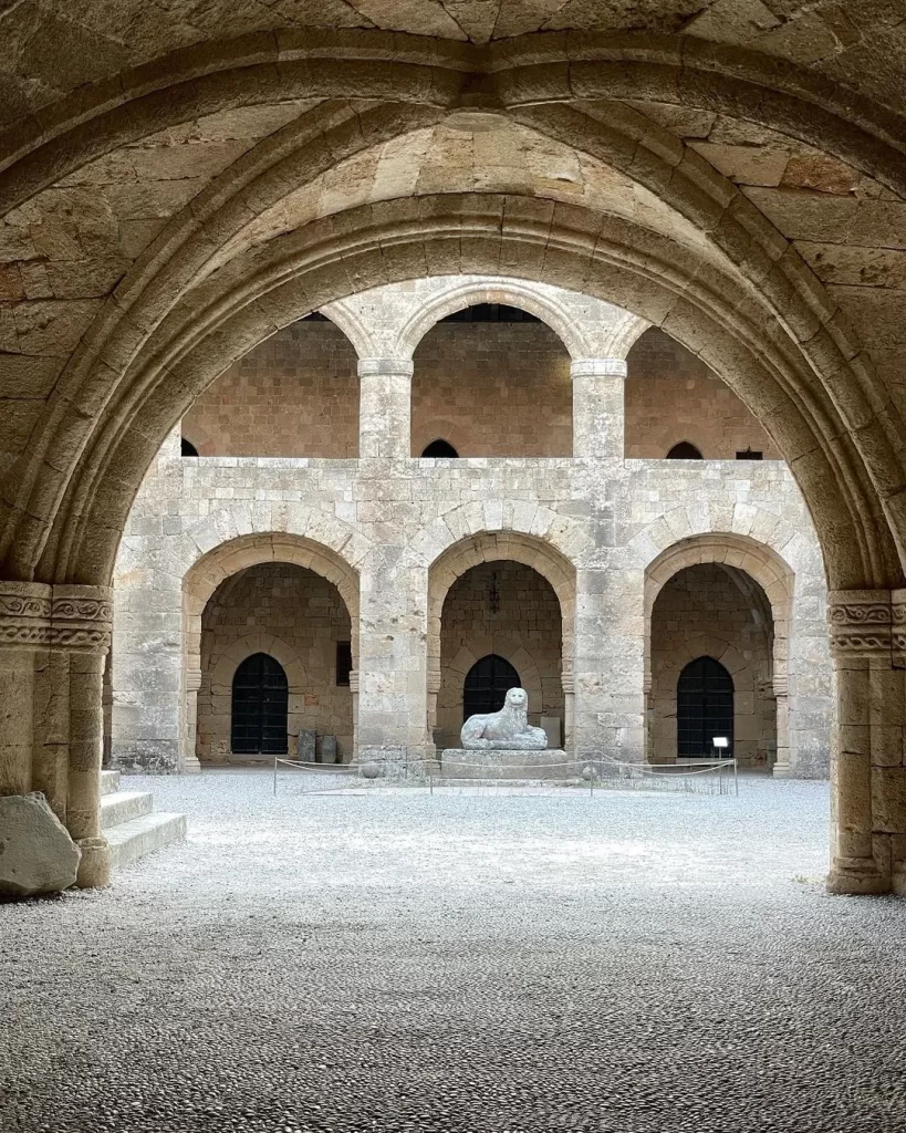 Rodos Arkeoloji Müzesi'nin etkileyici avlusunda, güçlü taş kemerlerin altında görünen gri çakıl taşlı zeminde duran, detaylı işlemeli büyük bir taş aslan heykeli, müzenin zengin tarihi koleksiyonunu ve ziyaretçilere sunulan eşsiz atmosferi yansıtıyor.