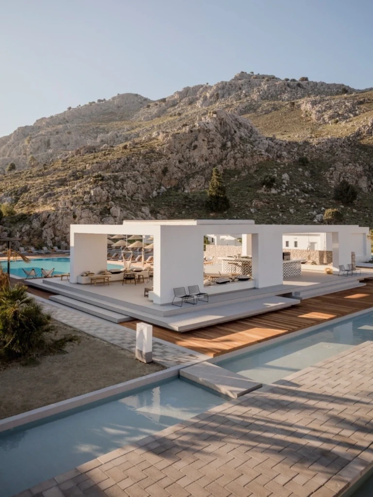 Rodos'ta gezilecek yerlerden biri olan Casa Cook otelinde, açık planlı modern beyaz yapılar, rahat oturma alanları, çevresindeki ahşap zeminler ve uzun havuzlarla tamamlanan, arka planda görkemli kayalık dağların yükseldiği dingin ve lüks bir atmosfer.