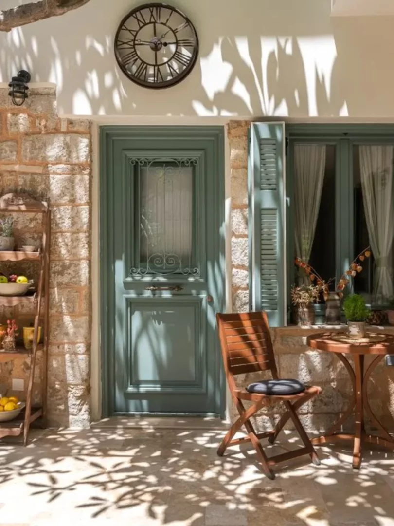 Rodos'ta gezilecek yerlerden biri olan Casa Natura'nın taş duvarlı, mint yeşili ahşap kapılı ve pencereli cephesinin önünde, duvarda büyük saatli, gölgeli avluda ahşap masa, sandalye ve dekoratif raflar güneş ışıkları altında huzurlu bir atmosfer sunuyor.