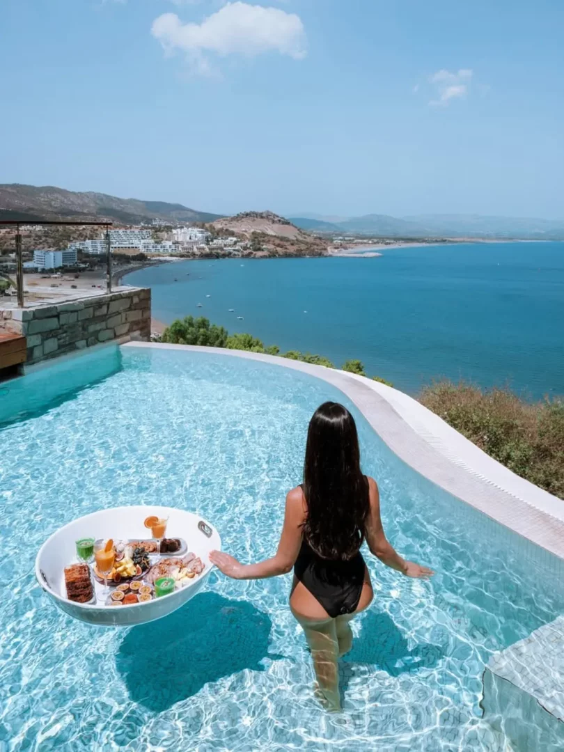 Rodos'taki Lindos Blu gibi lüks bir otelin sonsuzluk havuzunda, sırtı dönük, siyah mayolu uzun saçlı bir kadın, üzerinde taze meyveler, kahvaltılıklar ve içecekler bulunan beyaz yüzen bir servis tepsisini tutarak, Akdeniz'in masmavi sularına ve kıyıdaki yerleşimlere bakan eşsiz manzaranın tadını çıkarıyor.