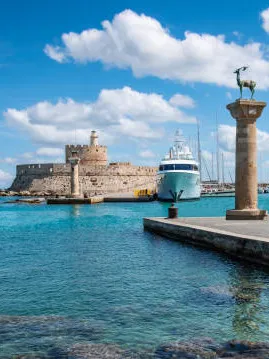 Rodos'un Mandraki Limanı'nda, berrak turkuaz denizin üzerinde süzülen beyaz bulutların gökyüzü altında, ucunda bronz geyik heykeli bulunan ikonik sütun ve modern bir yatın yanında uzanan tarihi Rodos Kalesi'nin görkemli surları görülüyor.