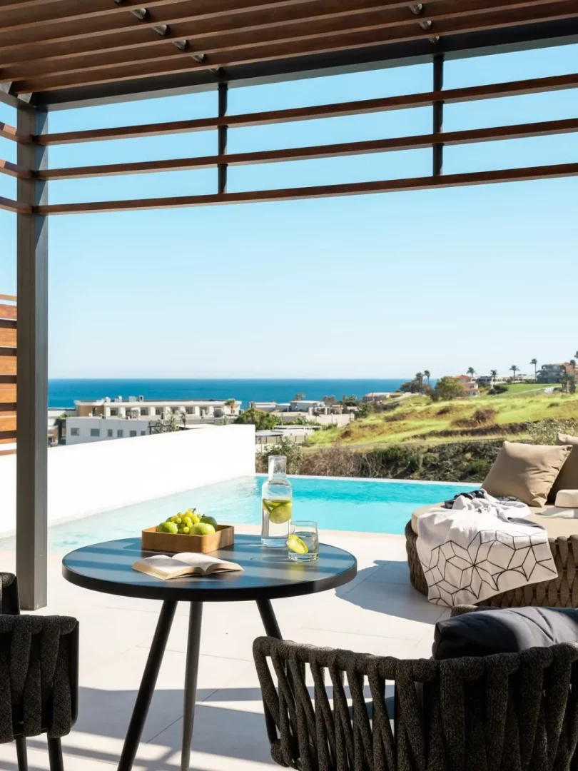 Rodos'taki Mitsis Alila Resort & Spa'nın şık balkonunda, Akdeniz'in masmavi sularına bakan özel havuzun yanında yer alan yuvarlak bir masada taze meyveler, limonlu su ve açık bir kitap bulunurken, huzurlu atmosferi yansıtan koyu renkli örgülü sandalyelerle çevrili bu lüks manzara, Rodos gezilecek yerler listesindeki konaklama seçeneklerinin kalitesini gözler önüne seriyor.