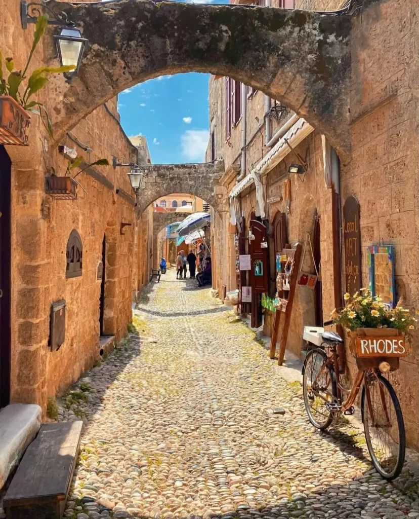 Rodos Eski Şehir'in dar, Arnavut kaldırımlı sokağında, üzerinde kemerli taş binaların yükseldiği, sağda çiçeklerle süslü RHODES yazılı bir bisikletin durduğu ve dükkanların sıralandığı, uzakta gezen insanların görüldüğü güneşli ve tarihi bir gezilecek yer.