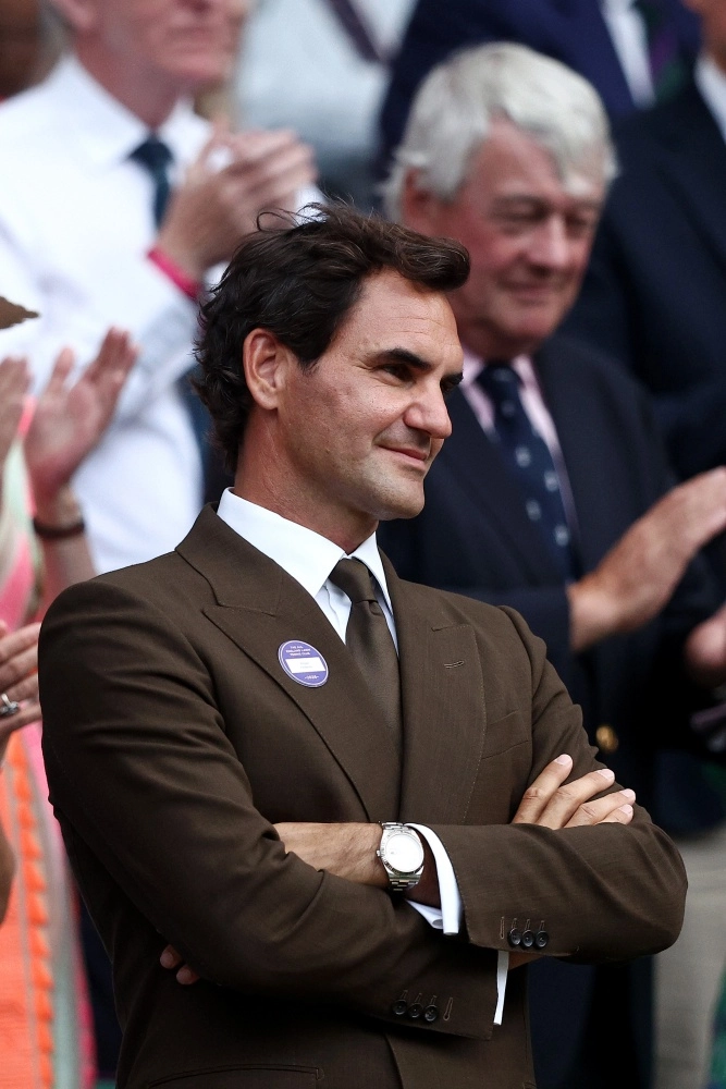 Roger Federer kahverengi bir takım elbise, beyaz gömlek ve kravatla, sol kolunda parlak bir Rolex Datejust kol saatiyle kolları bağlı bir şekilde hafifçe gülümseyerek Wimbledon'da seyircilerin arasında oturuyor.