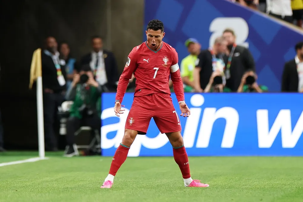 Portekiz'in 7 numaralı kırmızı formasıyla yeşil sahada, ağzı açık ve kolları hafifçe yana açılmış bir şekilde yoğun bir sevinç veya hırs ifadesiyle duran Cristiano Ronaldo, dikkat çekici pembe kramponlarıyla 2026 Dünya Kupası yolundaki kararlılığını simgeliyor.