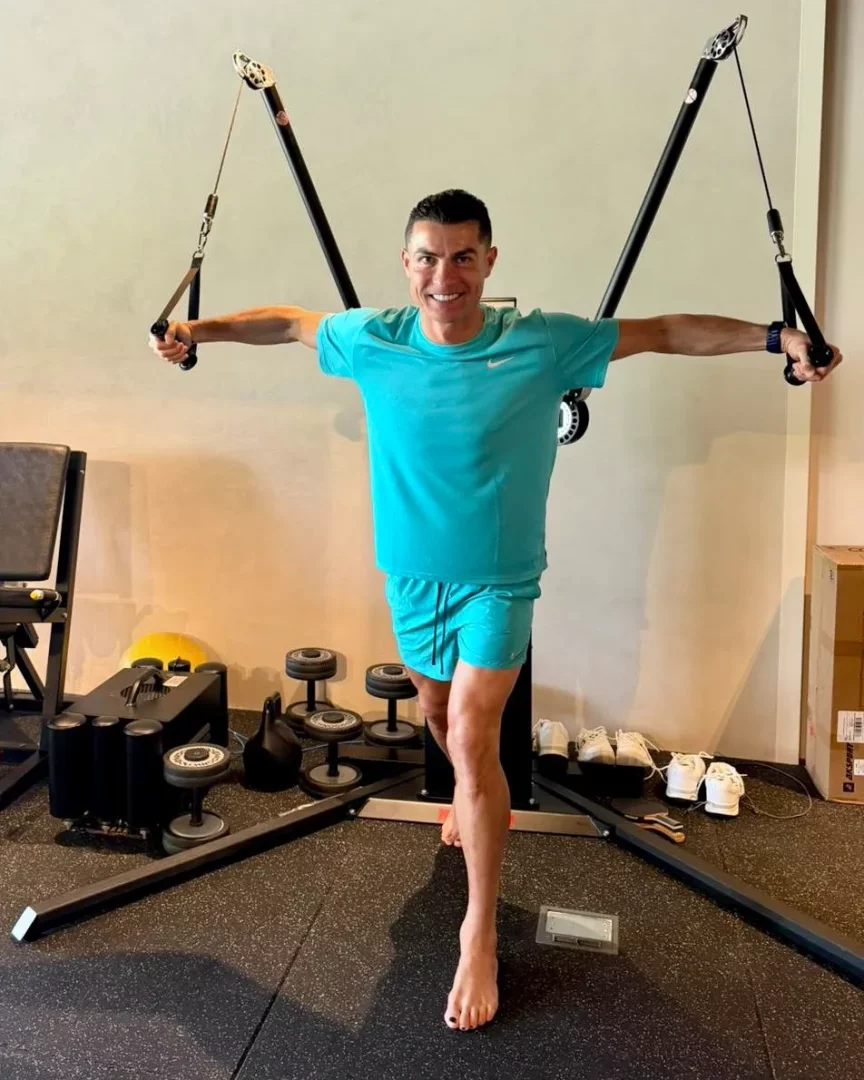 Ronaldo, formunu korumak ve futbol kariyerindeki uzun ömürlülüğünü sürdürmek amacıyla modern bir spor salonunda, kolları yana açık bir şekilde kablo makineleriyle dengeli bir egzersiz yaparak fit vücudunu ve enerjisini gözler önüne seriyor.