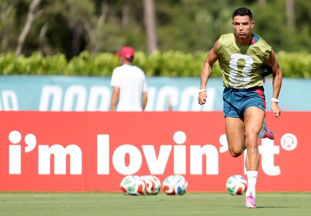 Cristiano Ronaldo, yeşil sahada sarı-yeşil antrenman yeleği ve lacivert şortuyla, kaslı vücudunu gösteren yoğun bir egzersiz sırasında kararlı bir ifadeyle hızla koşuyor; bu görüntü, futbol kariyerindeki uzun solukluluğunun sırrını vurguluyor.