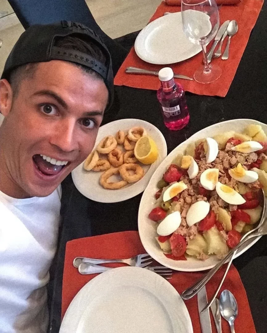 Cristiano Ronaldo, arkasında siyah şapka ve beyaz tişörtüyle, masasında bol yumurtalı, ton balıklı ve sebzeli salata ile kalamar halkaları ve pembe bir içecek bulunan bir öğünle selfie çekerken, sağlıklı beslenmenin spor kariyerindeki önemini vurguluyor.