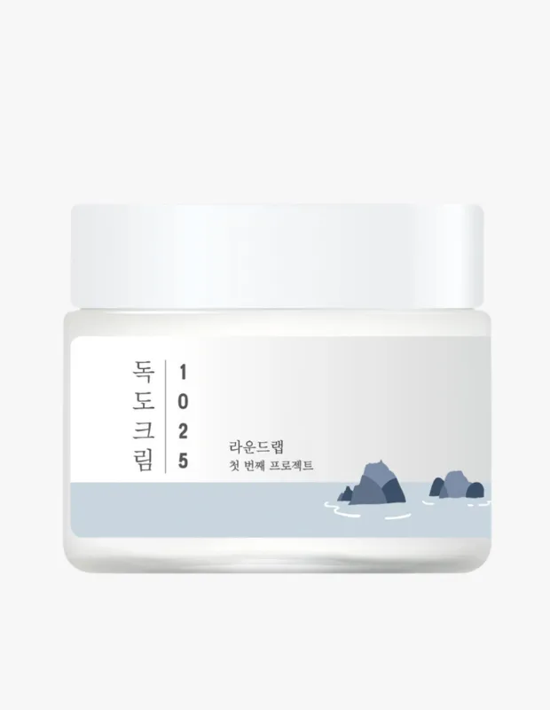 Sade tasarımlı beyaz kapaklı Round Lab 1025 Dokdo Kremi kavanozu; üzerinde Korece Dokdo 크림 yazısı, 1025 sayıları ve markayı belirten etiketinde, açık mavi su üzerinde koyu gri kayalık adaların minimalist bir çizimi yer alıyor.