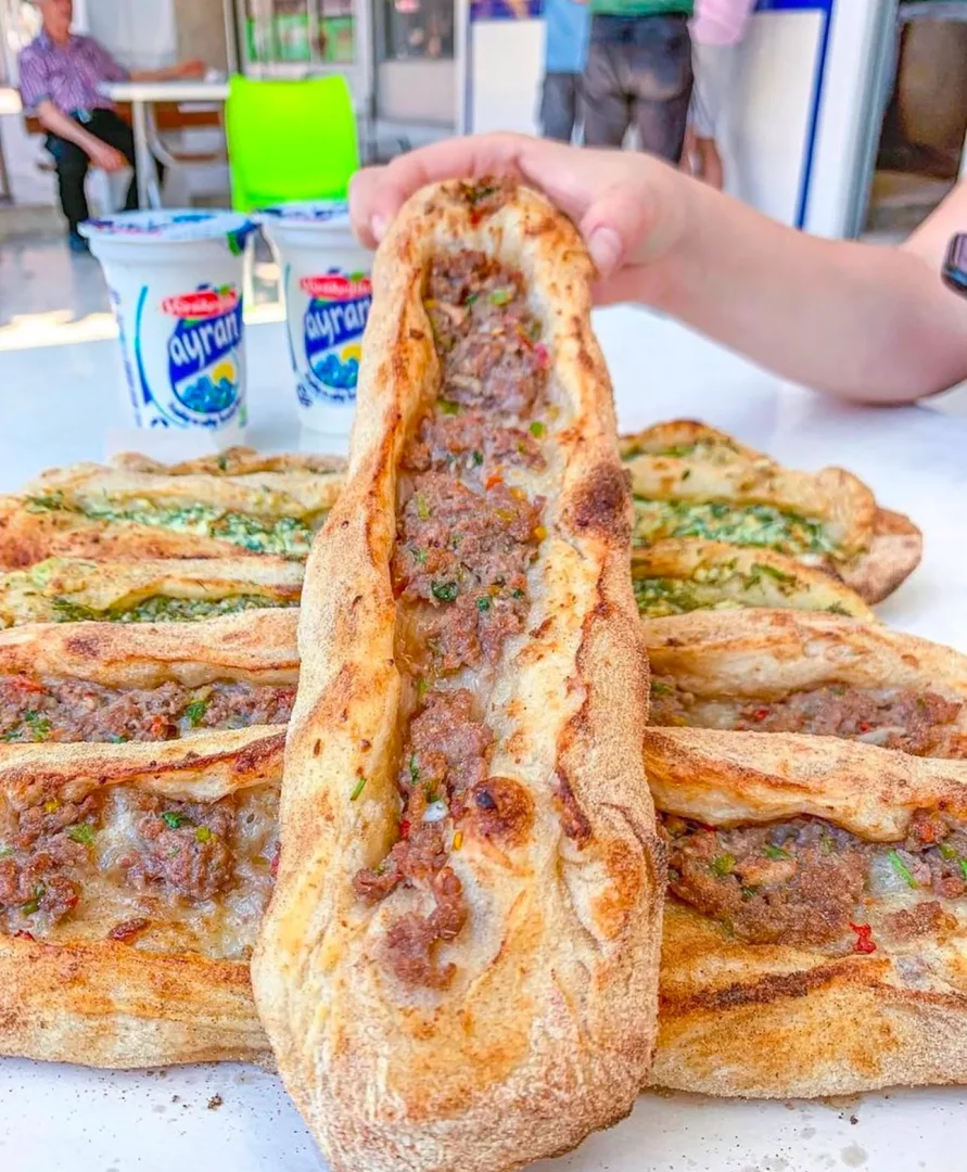 Bir el, geleneksel Sakarya pidesi gibi görünen, kıyma dolgulu, uzun ve sıcak bir pideyi özenle tutarken, masada başka lezzetli pideler ve yanında serinletici ayranlar bu fırın atmosferini tamamlıyor.