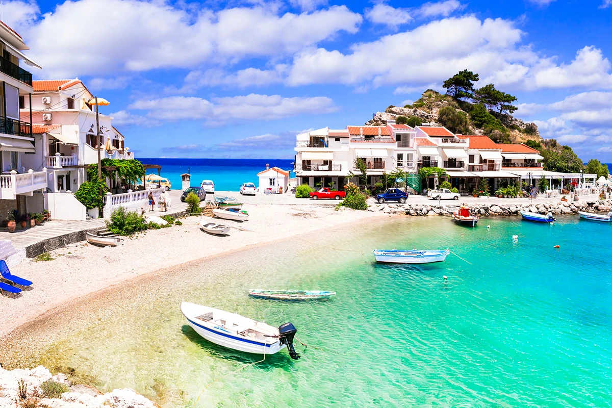 Samos Adası'nda, beyaz binaların ve kırmızı çatılı evlerin çevrelediği turkuaz renkli denizde demirli teknelerle dolu pitoresk bir koy, adaya nasıl gidileceğini anlatan yazının görselini oluşturuyor.