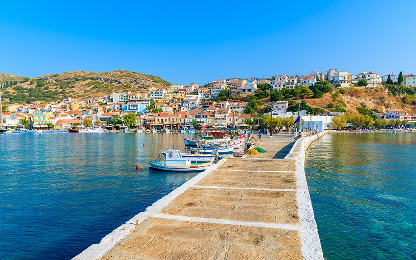 Samos'ta kapıda vize kolaylığıyla erişilebilen, güneşli bir günde berrak turkuaz sularında demirlemiş rengarenk balıkçı tekneleri ve yemyeşil tepelerin eteklerine sıralanmış pastel tonlu evleriyle şirin bir sahil kasabası manzarası.