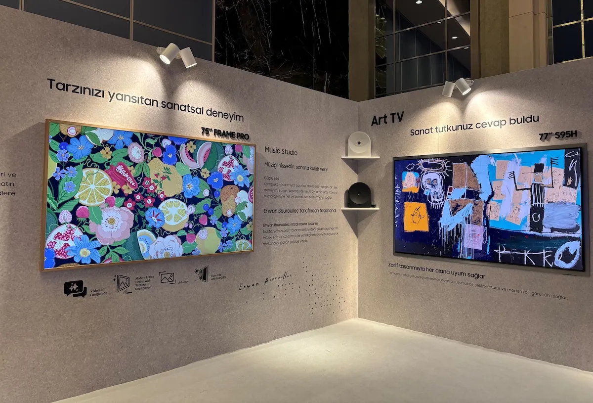 Parlak ışıklarla aydınlatılmış gri bir duvarda sergilenen, sanat eseri gibi duran iki Samsung yapay zeka TV: soldaki çerçeveli model canlı çiçek ve meyve desenleri gösterirken, sağdaki model soyut bir tabloyu yansıtarak Samsung'un 2026 AI TV serisinin estetik ve teknolojik yeniliklerini gözler önüne seriyor.