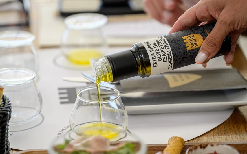 Bir elin, üzerinde Santo Roots logosu ve Extra Virgin Olive Oil ibaresi bulunan koyu renkli bir şişeden altın sarısı zeytinyağını şeffaf bir tadım bardağına özenle akıtırken, Santorini zeytinyağı tadım etkinliğinin davetkar masası dikkat çekiyor.