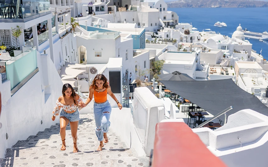 İki genç kadın, Santorini'nin karakteristik beyaz badanalı binaları ve Ege Denizi manzarası eşliğinde, taş döşeli merdivenlerden neşeyle iniyor, bu an gezilecek yerleri keşfetmenin keyifli bir örneğini sunuyor.