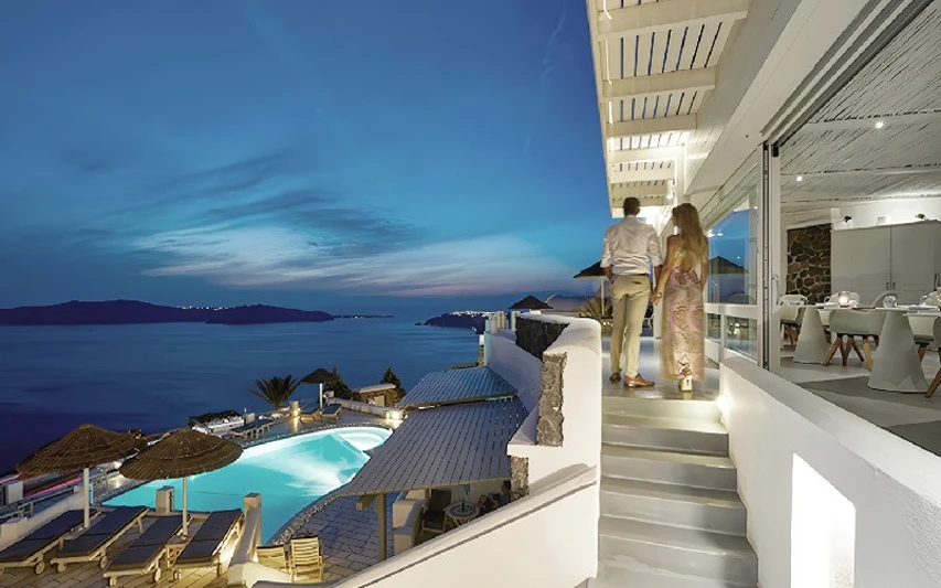 Alacakaranlıkta Ege Denizi'ne nazır bir Santorini spa otelinde, romantik bir akşam yemeği öncesi havuz ve deniz manzarasının keyfini çıkaran bir çift, beyaz duvarlı merdivenlerden aşağıya doğru el ele yürüyor.