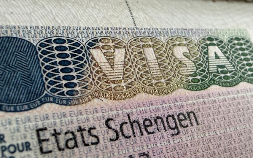 Yakın çekimde bir pasaport sayfasında bulunan, güvenlik özellikleriyle dikkat çeken, VISA kelimesinin altın ve yeşil tonlarında parladığı bir Schengen vizesi etiketi görülüyor; bu görsel, Santorini seyahati için vize gereklilikleri hakkında bilgi veren makaleye kapıda vize süreçleriyle ilgili önemli bir bağlam sunuyor.