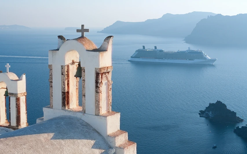 Santorini'ye nasıl gidileceğini düşündüren bu görselde, adanın simgesi haline gelmiş beyaz çan kuleleri ön plandayken, masmavi Ege Denizi üzerinde ilerleyen büyük bir yolcu gemisi adaya ulaşım seçeneklerinden birini gösteriyor ve arka planda volkanik dağlar ufukta kayboluyor.
