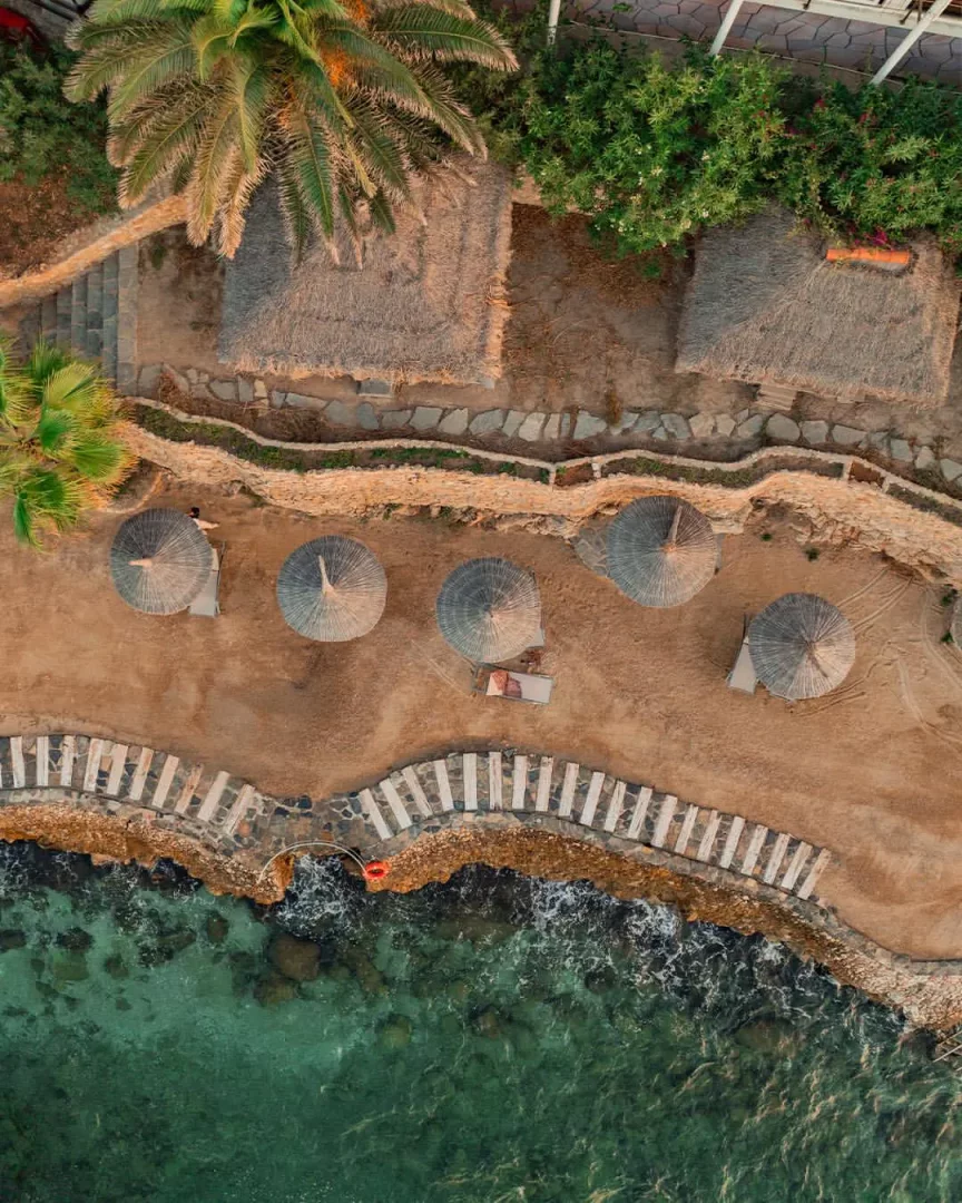 Sarpedor Boutique Hotel'in Torba Plajı'ndaki lüks atmosferini yansıtan bu havadan görünümde, altın rengi kumsalda yer alan bej hasır şemsiyelerin altında uzanan şezlonglar, kıyı şeridini çevreleyen turkuaz sular ve palmiye ağaçları arasındaki saz çatılı butik yapılar doğal güzelliği sergiliyor.