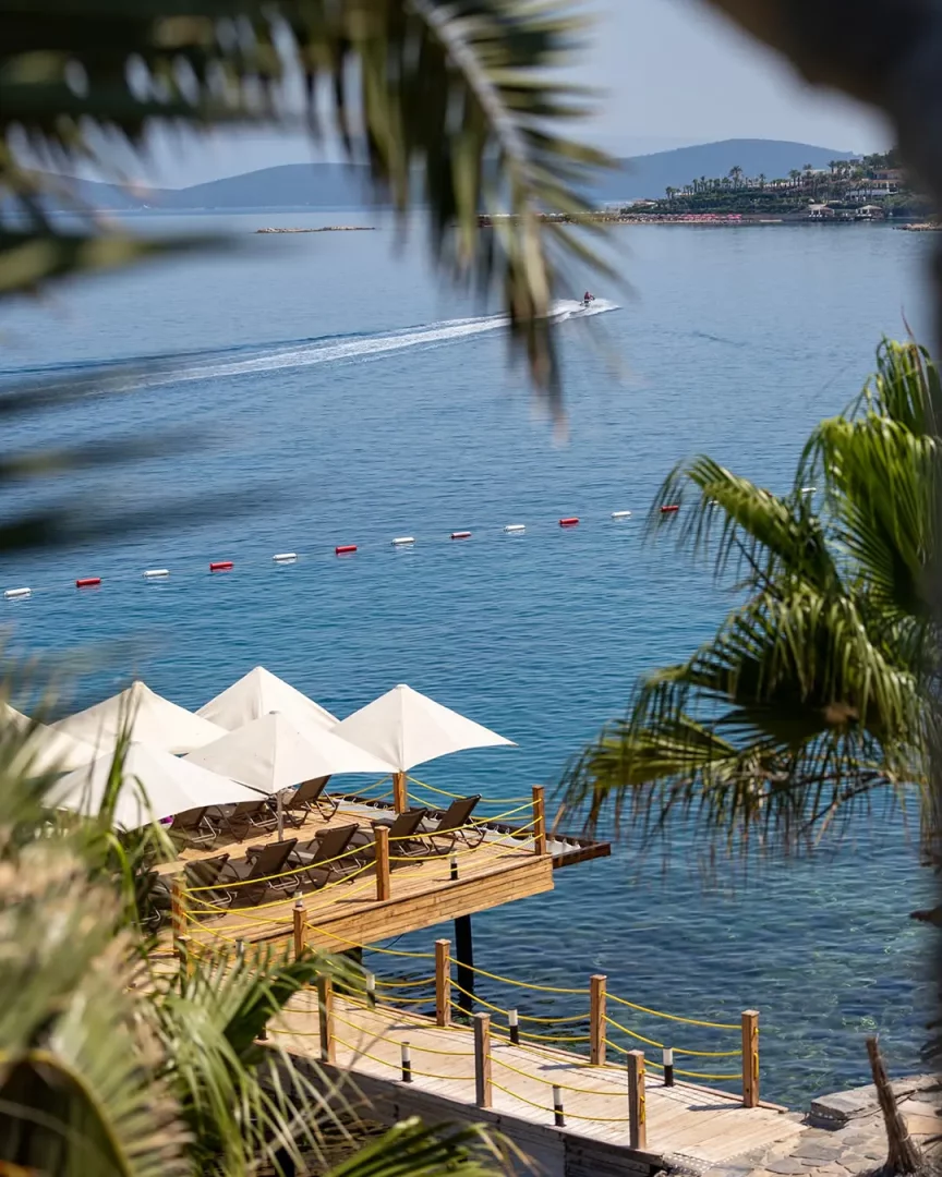 Sarpedor Boutique Hotel'in Torba kıyısındaki ahşap iskelesi, şık şezlonglar ve beyaz şemsiyelerle donatılmış güneşlenme platformuyla berrak mavi denize uzanırken, uzakta hızla giden bir jet ski su yüzeyinde iz bırakıyor.