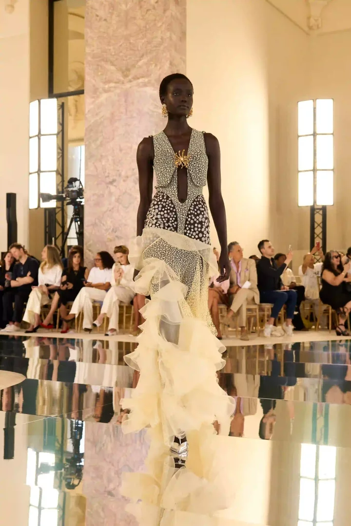 Bir model, derin V yakalı, altın yıldız broşlu, benekli üst kısmı ve volümlü bej fırfırlarla süslü heykelsi bir haute couture elbiseyle Schiaparelli Sonbahar-Kış 2026 defilesinde podyumda yürürken, yansıtıcı zeminde izleyiciler ve kendisinin yansıması görülüyor.