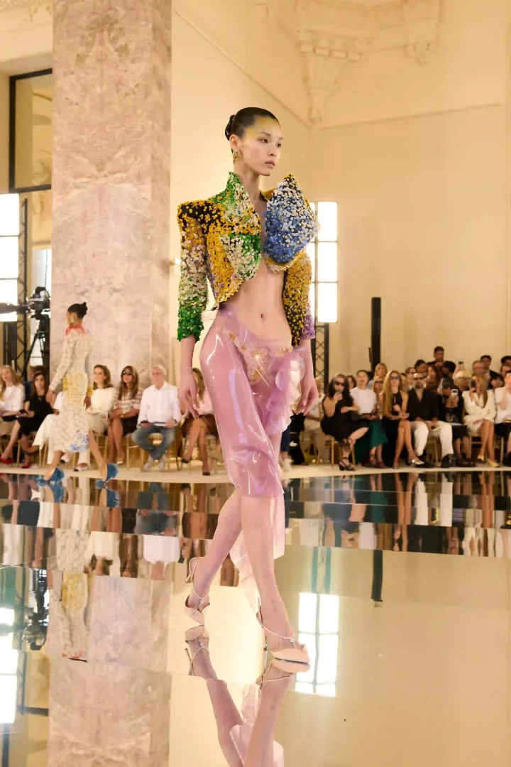 Paris Haute Couture Moda Haftası'nın dikkat çeken anlarından birinde, Schiaparelli Sonbahar-Kış 2026 koleksiyonundan bir model, omuzları heykelsi ve yoğun renkli işlemeli kısa bir ceket ile saydam, açık pembe lateks şort giyerek yansıtıcı podyumda yürüyor, koleksiyonun yenilikçi malzemeler ve sürreal silüetlerle couture dünyasındaki sınırları zorlayan yaklaşımını sergiliyor.