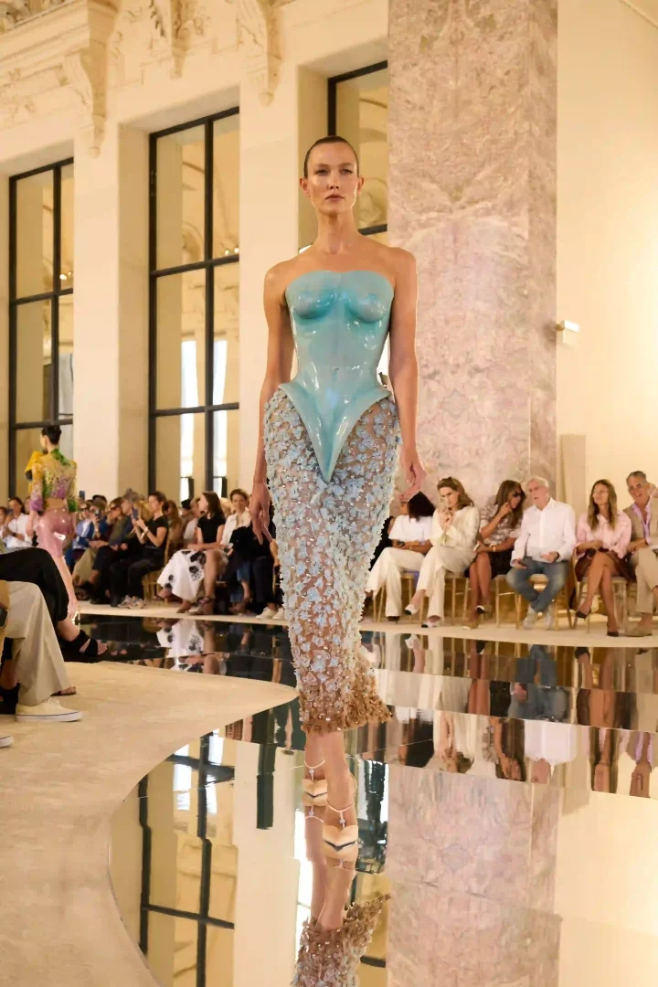 Bir model, Paris Haute Couture Moda Haftası'nda Schiaparelli'nin Sonbahar-Kış 2026 koleksiyonundan, vücut hatlarını vurgulayan parlak turkuaz lateks büstiyeri ve uçuk mavi çiçek işlemelerinin etek ucuna doğru kahverengiye dönüştüğü heykelsi şeffaf elbisesiyle podyumda yürüyor.