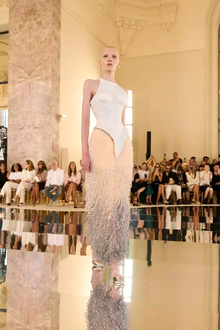 Schiaparelli Sonbahar-Kış 2026 haute couture defilesinde aynalı podyumda ilerleyen model, heykelsi beyaz büstiyer ile şeftali tonlarından gümüşe geçiş yapan yoğun saçaklı eteğiyle Daniel Roseberry'nin sürrealist vizyonunu gözler önüne seriyor.
