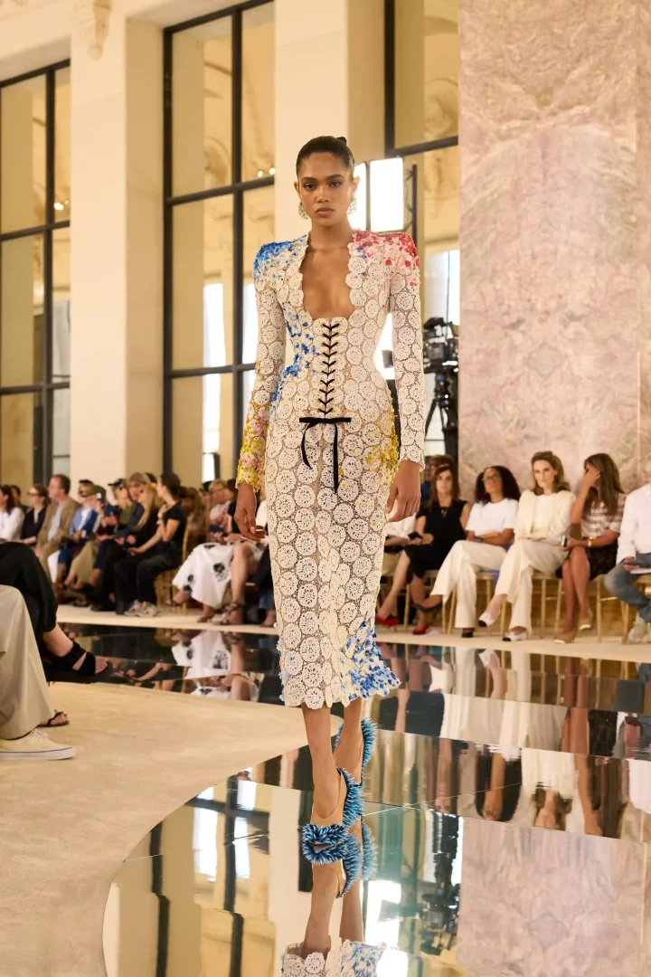 Model, Schiaparelli Sonbahar-Kış 2026 Haute Couture koleksiyonundan, yaratıcılığın ve malzeme deneylerinin sınırlarını zorlayan, derin V yakalı, önde siyah bağcıklı korseli, beyaz dairesel dantel motifleriyle bezenmiş, omuzlarında ve etek uçlarında canlı mavi, kırmızı, sarı renk sıçramaları olan uzun kollu midi bir elbise içinde yansımalı podyumda yürüyor.