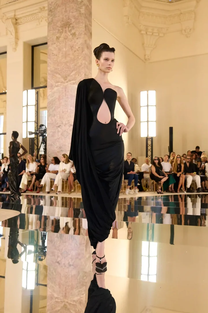 Yansıtıcı bir podyumda, Schiaparelli Sonbahar-Kış 2026 Haute Couture koleksiyonundan, asimetrik kesimli, göğüs ve karın bölgesinde oval bir penceresi olan, bir omzu drape detaylı uzun kollu siyah bir elbise giymiş model; Daniel Roseberry'nin yaratıcılığın kalıplara sığmadığını vurgulayan deneysel haute couture siluetlerini sergiliyor.
