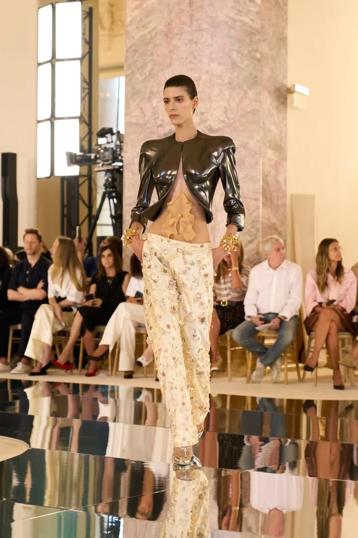 Schiaparelli Sonbahar-Kış 2026 Haute Couture koleksiyonundan, parlak koyu renkli kısa ceketi, ten rengi organik silikon büstiyeri ve inci işlemeli krem rengi pantolonuyla aynalı podyumda yürüyen bir manken, Daniel Roseberry'nin yaratıcılığın sınırlarını zorlayan yeni siluetlerini ve deneysel malzeme kullanımını vurguluyor.