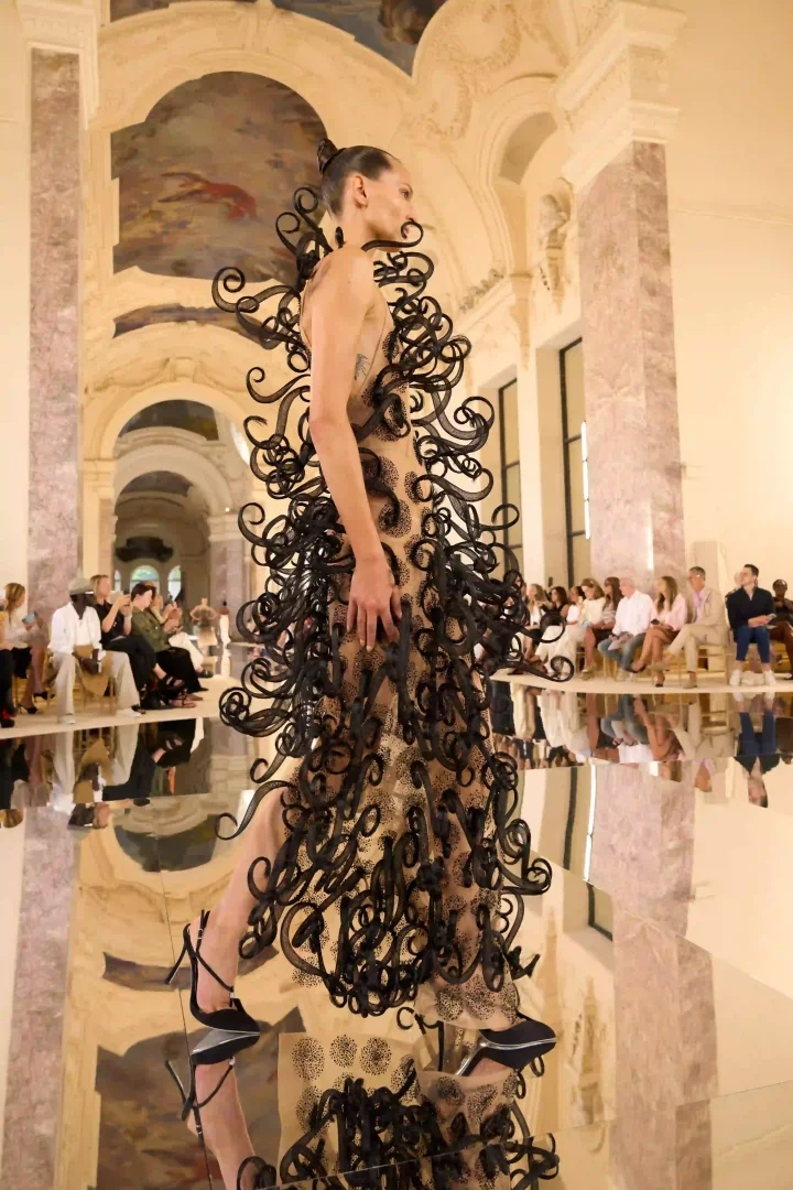Paris Haute Couture Moda Haftası kapsamında, Schiaparelli Sonbahar-Kış 2026 defilesinden, Gaudí'nin organik mimarisinden ilhamla yaratılmış, ten rengi transparan kumaş üzerine işlenmiş siyah kıvrımlı, heykelsi ve kinetik dokunaçları andıran detaylarla süslü bir elbiseyi taşıyan model aynalı podyumda yürürken görülüyor.