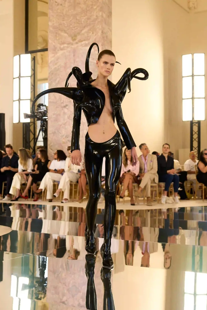Schiaparelli Sonbahar-Kış 2026 Haute Couture koleksiyonundan, parlak siyah lateks ve heykelsi detaylarla omuzları çevrili, kinetik dokunuşlu bir ceket ile uyumlu dar pantolon giyen bir model, yansıtıcı podyumda güçlü bir duruş sergiliyor.
