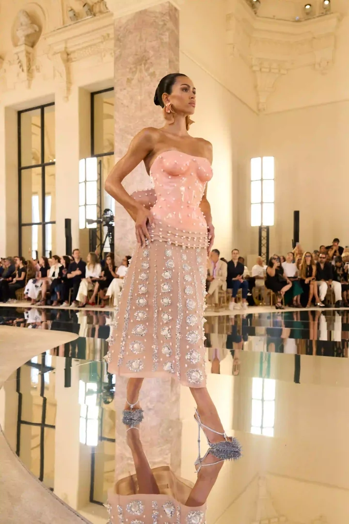 Bir model, Paris Haute Couture Moda Haftası'nda tanıtılan Schiaparelli Sonbahar-Kış 2026 koleksiyonundan, hipergerçekçi büstiyerli, etek kısmında parlak gümüş ve beyaz süslemelerle işlenmiş heykelsi pembe straplez elbisesi ve aynı tonda büyük küpeleriyle yansımalı podyumda yürüyor.