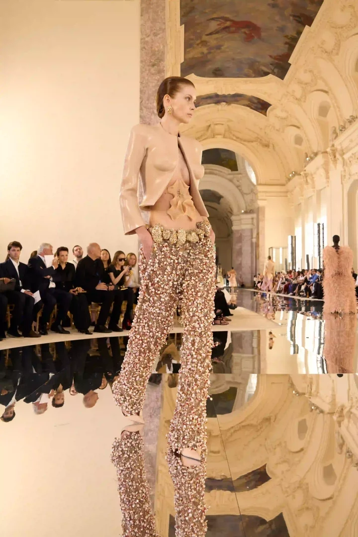 Paris Haute Couture Moda Haftası'nda Schiaparelli Sonbahar-Kış 2026 defilesinden, lateks benzeri bej bir üst ile göbeğindeki heykelsi silikon detayı ve tamamı minik, parıltılı süslemelerle kaplı, yüksek belli pantolon giyen bir model, görkemli bir salondaki yansıtıcı podyumda yürüyerek haute couture'ün iddialı estetiğini sergiliyor.