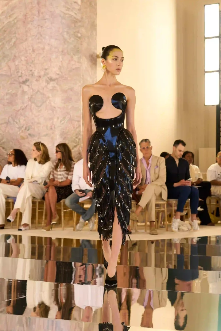 Paris Haute Couture Moda Haftası'nın dikkat çeken anlarından Schiaparelli Sonbahar-Kış 2026 koleksiyonuna ait bir görselde, heykelsi, parlak siyah ve askısız bir lateks elbise giyen, göz kapakları sarı makyajlı bir model, yansımalı podyumda izleyicilerin önünde kararlı adımlarla yürüyor; elbisenin göğüs kısmındaki belirgin dairesel detaylar ve etek kısmındaki metal işlemeli katmanlar dikkat çekiyor.