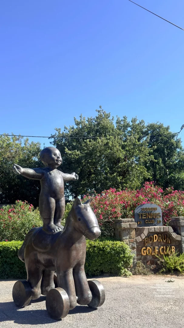 Bodrum Golf Kulübü tabelasının önünde, kollarını iki yana açmış, tekerlekli at üzerindeki bronz bebek heykeli, yemyeşil ağaçlar ve pembe çiçekli çalılarla çevrili açık havada sergileniyor.