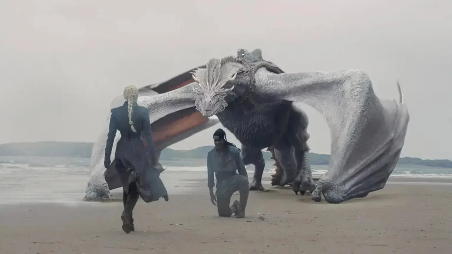 House of the Dragon dizisinden Seasmoke adlı, geniş kanatları açık büyük açık gri ejderhanın bulutlu bir gökyüzü altında kumlu bir plajda bulunduğu, örgülü sarı saçlı bir kadının ona doğru yürüdüğü ve koyu tenli bir adamın önünde diz çöktüğü bir sahne.